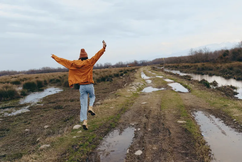 Woman lifestyle outdoors hiking on muddy path in winter landscape, arms raised in joy, near river stream, open countryside fields, calm nature under clouds, bright sunny scenic trail