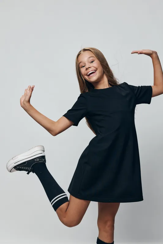 Girl posing in black dress with striped socks and sneakers jumping on gray background. Cute smiling child with long hair in casual clothing with energetic friendly expression.