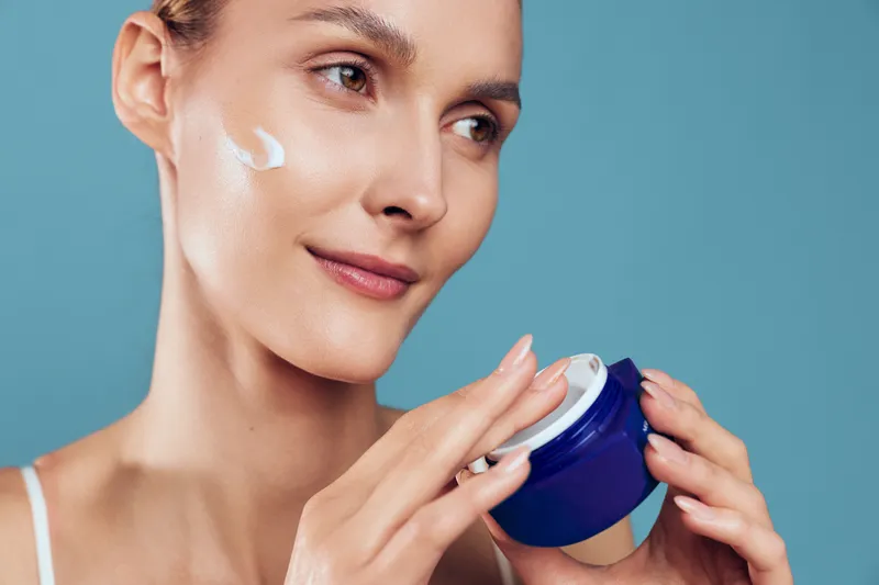 Woman applies skincare cream from blue jar to her face closeup, gentle smile, beauty routine concept, spa skincare moment, isolated on blue background, healthy glow