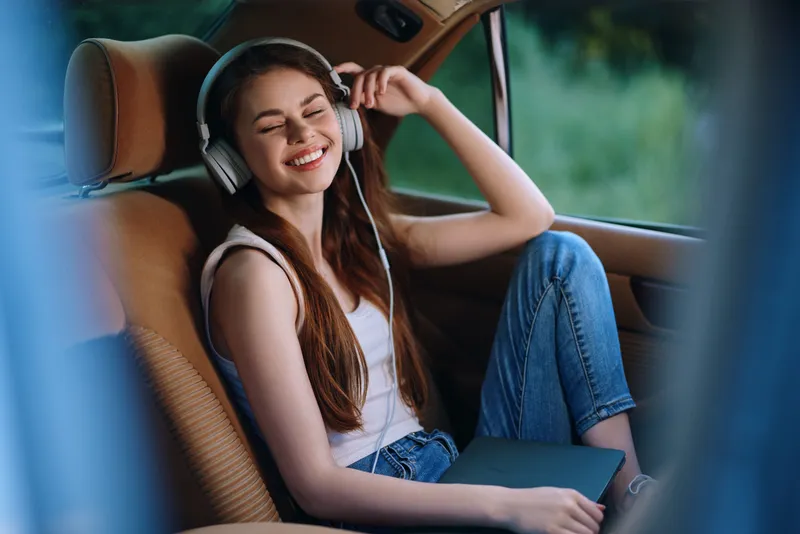 Young woman enjoying music in a car, wearing casual clothes and headphones, reflecting happiness and relaxation in a sunny setting.