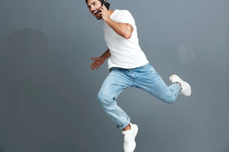 Joyful man jumping while talking on mobile phone, wearing casual white t-shirt and blue jeans, isolated on solid gray background. Energetic lifestyle concept.