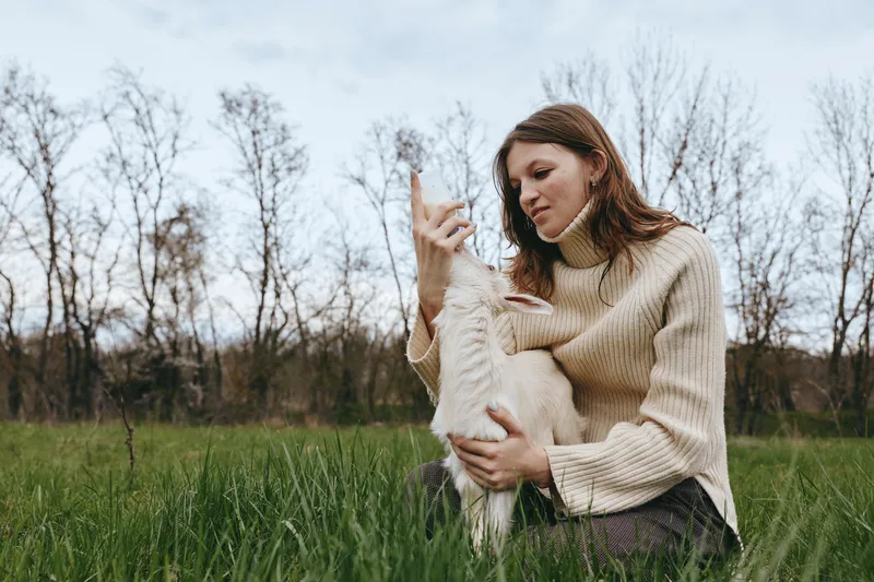 Woman in a cozy sweater sits in a green meadow, embracing a fluffy goat kid, sharing gentle affection outdoors, conveying wholesome lifestyle moments and natural bond with animals.