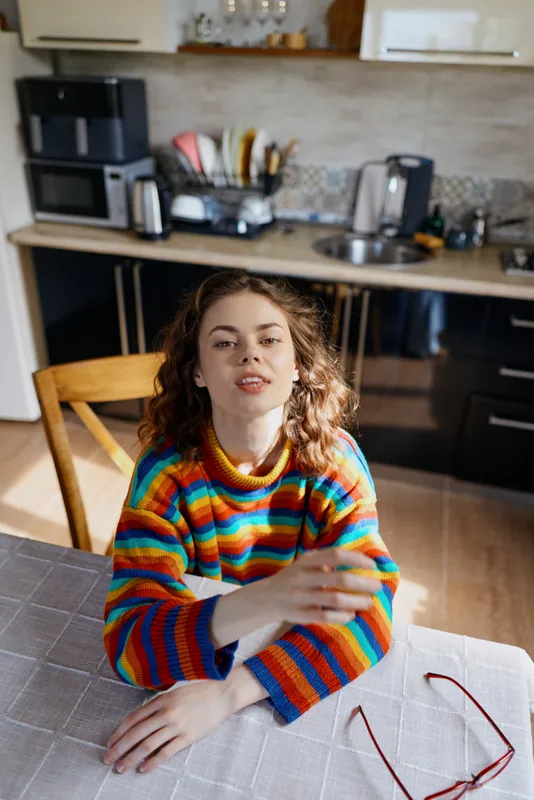 Young woman with curly brown hair sits at a table in a bright kitchen wearing a colorful striped sweater, looking playfully at the camera.