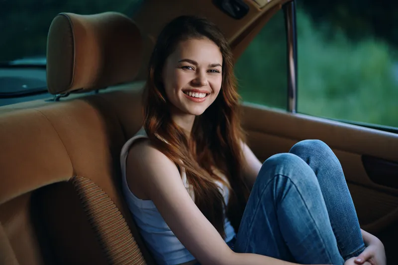 Young woman with long hair smiling in the backseat of a car, wearing casual attire and enjoying a sunny day, capturing a joyful moment.