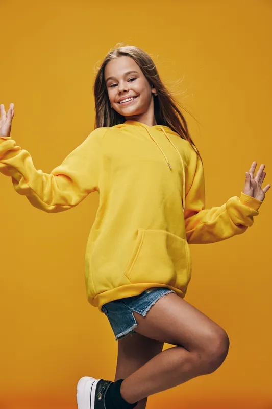 Teenage girl smiling and dancing in yellow hoodie and denim shorts on orange background. Happy young female enjoying music with closed eyes and carefree feeling in studio.