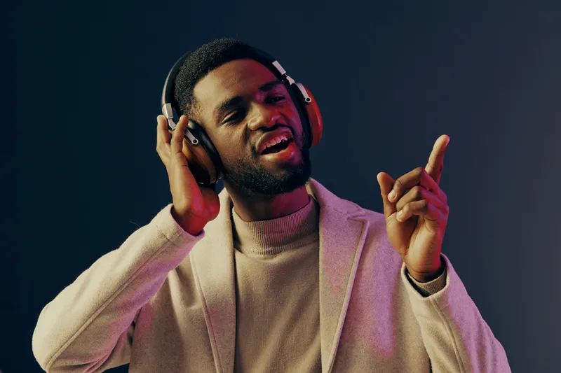 Happy young Black man with headphones enjoying music in a modern setting, wearing a stylish sweater and a coat against a colorful gradient background