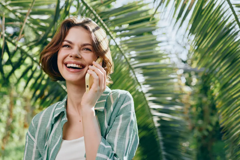 Bright portrait of a smiling woman outdoors, sunlit garden with palm leaves, wearing a casual striped shirt and radiant mood.