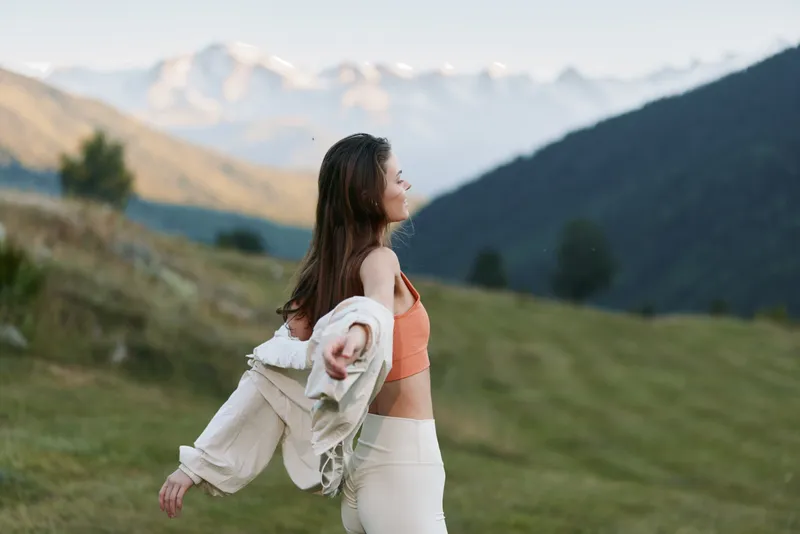 A cheerful woman stands outdoors in a scenic landscape, smiling as she sheds a light jacket, enjoying fresh air and sunshine in a peaceful countryside setting.