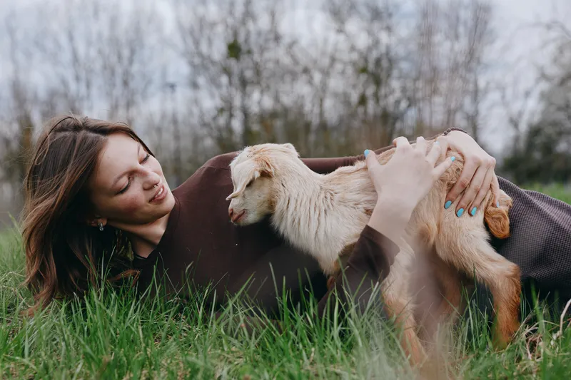 A woman lies on grass beneath a warm blanket, cradling a goat kid in a bright outdoor scene. The moment captures calm bonding, rural living, and affection between human and animal.