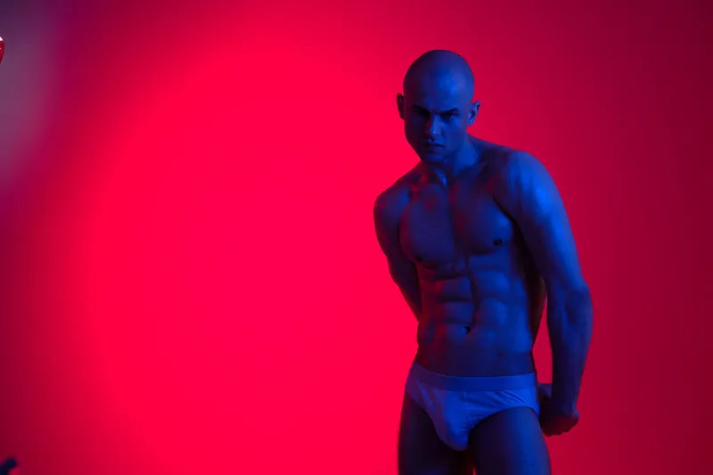 Strong man with toned physique poses dramatically against a vibrant gradient background, showcasing powerful lighting and shadows for an impactful visual effect.