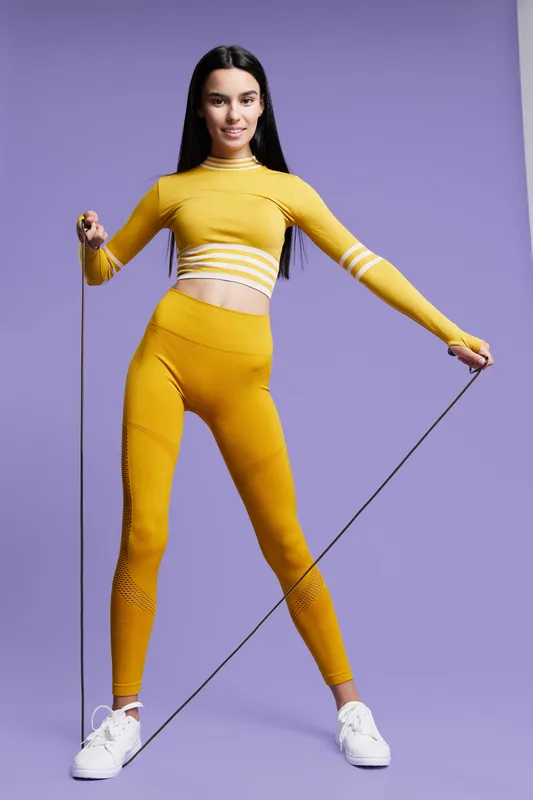 Woman sport fitness wearing yellow activewear jumps with a skipping rope against purple background, showing energy, health, and exercise motivation indoors.