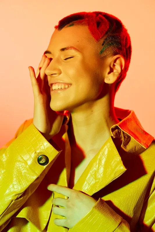 Smiling woman with short hair wearing a bright yellow textured jacket, expressing joy and warmth against a vibrant orange background, showcasing modern fashion.