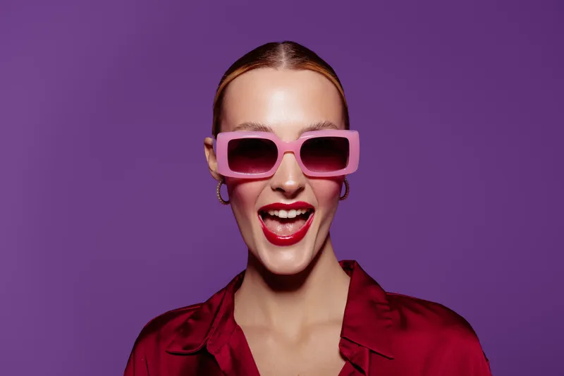 Happy woman with pink sunglasses and red lipstick smiles widely against purple background. Fashion portrait with bright colors and confident expression. Concept of style and joy.