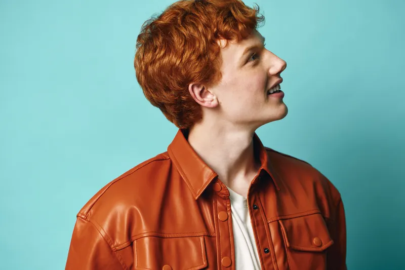 Young man with red hair wearing brown leather jacket looking sidewards on blue background. Casual style, portrait and fashion concept with confident expression and trendy outfit.