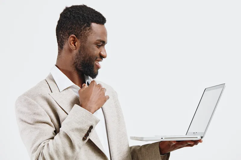 Smiling man in beige blazer holding laptop in one hand and touching chin with other, looking at screen, clean background, professional attire, business concept, technology, workspace.