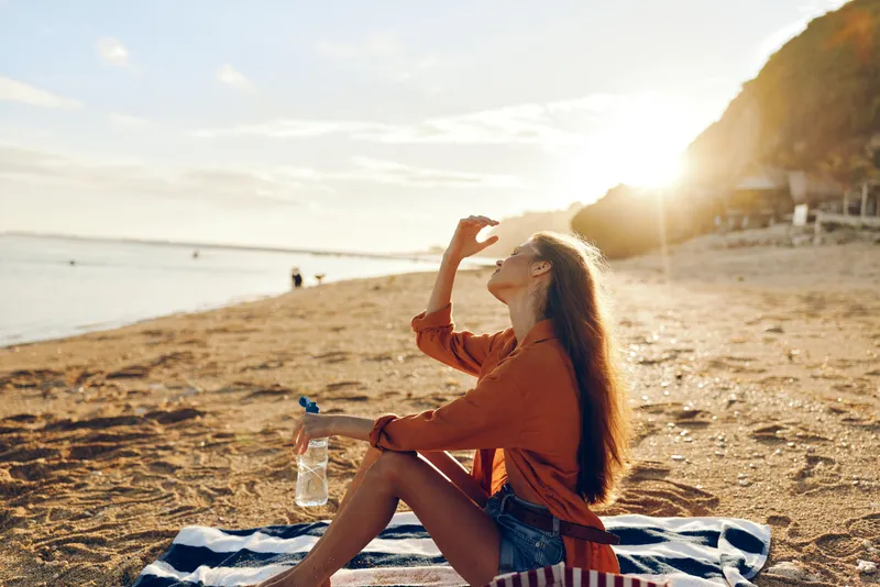 Woman at beach sunset, relaxing on striped blanket, sipping water from bottle, hand in hair, warm sunlight, calm ocean horizon, casual travel mood, golden color tone, soft breeze, gentle waves