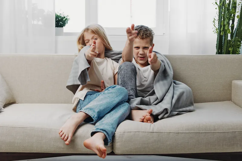 Two playful children sitting on sofa covered with blanket pretending to shoot fingers, enjoying cozy home moment together.