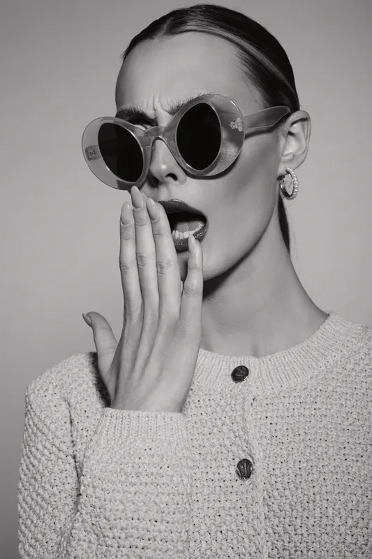 Young white woman with light skin in round sunglasses covers mouth with hand, showing surprise in black and white portrait photography.