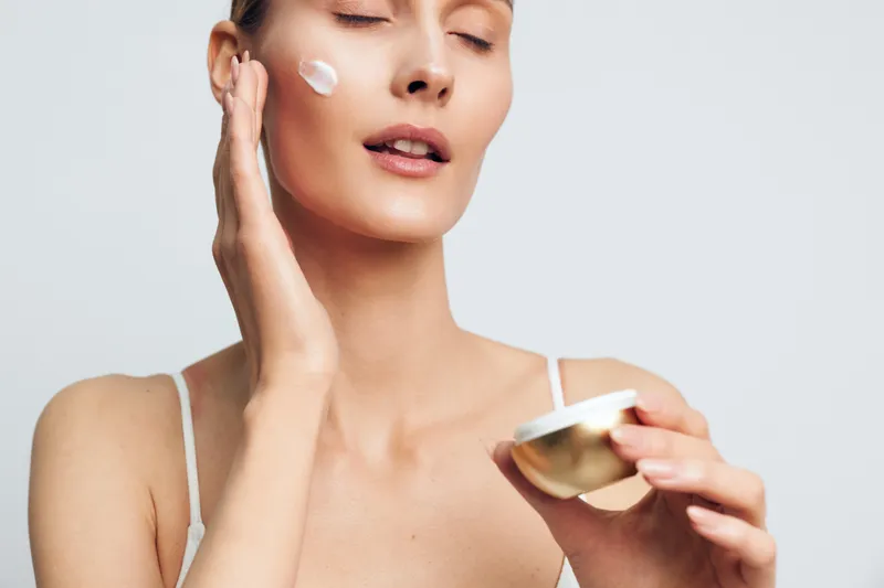 Young woman applying face cream with closed eyes holding container in hand. Skincare routine concept with soft natural lighting on white background for beauty and wellness.