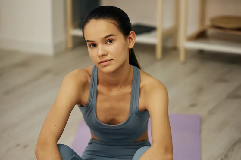 Young woman fitness portrait in athletic sportswear sitting on a yoga mat indoors, showing a relaxed pose after workout with a focused expression and minimal home gym background.