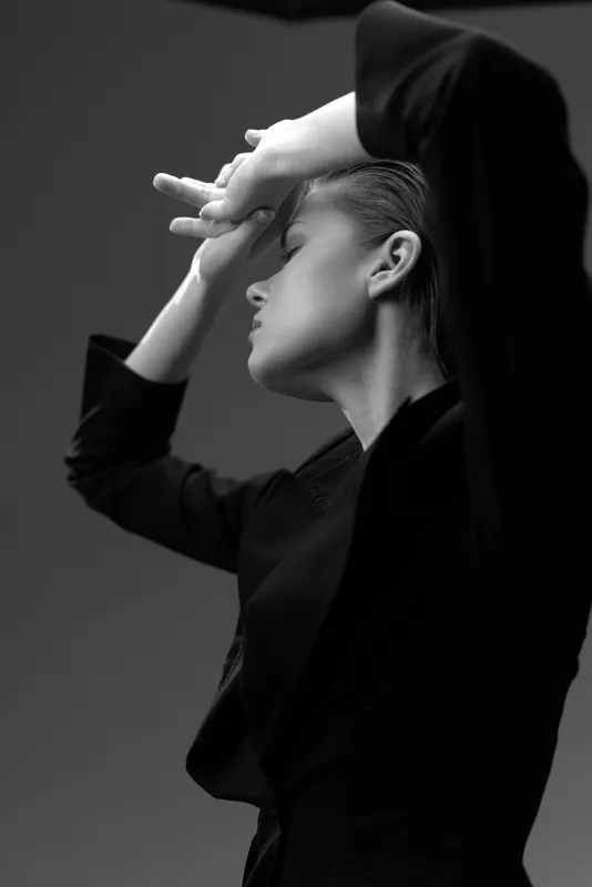 Black and white portrait of a thoughtful woman with closed eyes and raised arm, wearing dark clothing in a minimalistic setting.