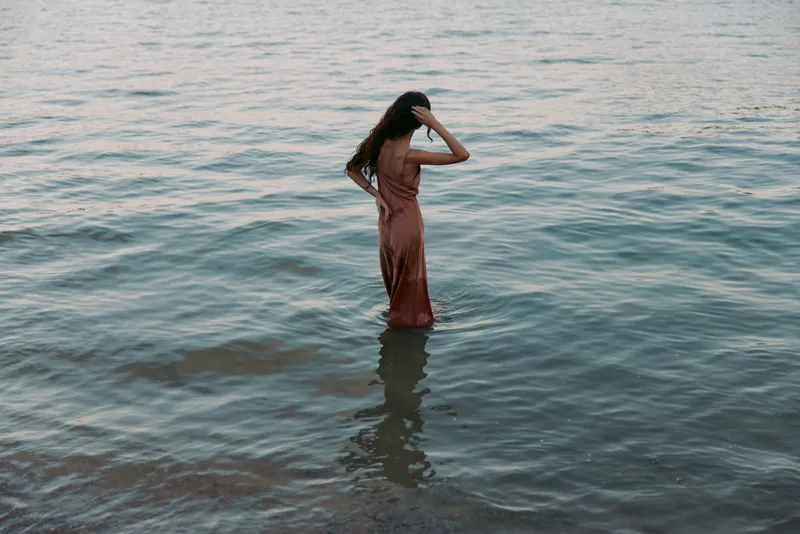 A lone woman stands waist-deep in calm water, wearing a flowing long dress, hair loose as soft light glints on the surface, creating a serene, contemplative mood.