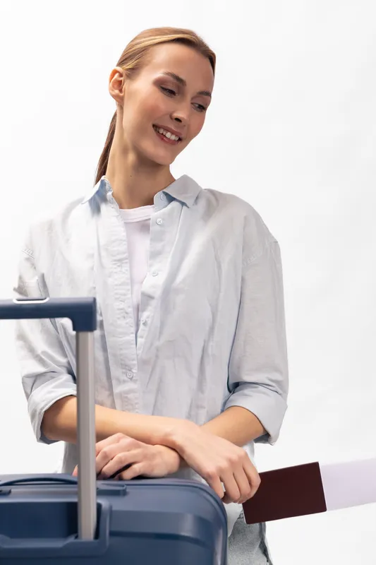 Smiling woman in casual light shirt holding passport and suitcase handle, looking down. Concept of travel, vacation, and female traveler with positive mood and light background.