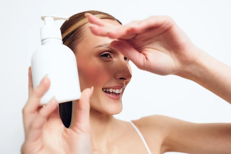 A smiling woman uses a pump bottle for skincare, applying product to her face in a bright bathroom. Clear focus on self care, beauty routine, and healthy skin.