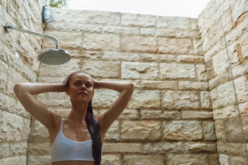 woman showering outdoors under rain showerhead with stone wall background, wet hair, relaxed mood, natural light, summer wellness concept