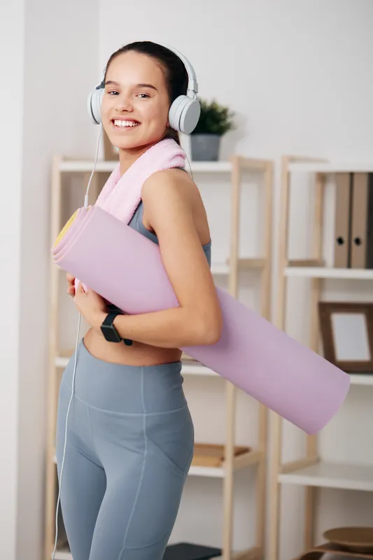 Fitness woman in a gym environment, holding a pastel purple yoga mat, wearing headphones and a light towel, smiling during a focused workout session.