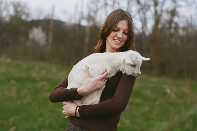Joyful woman outdoors, holding a goat kid, soft sunlight, gentle breeze, casual clothing, warm smile, rural setting, fresh grass, peaceful moment, wholesome lifestyle, candid connection