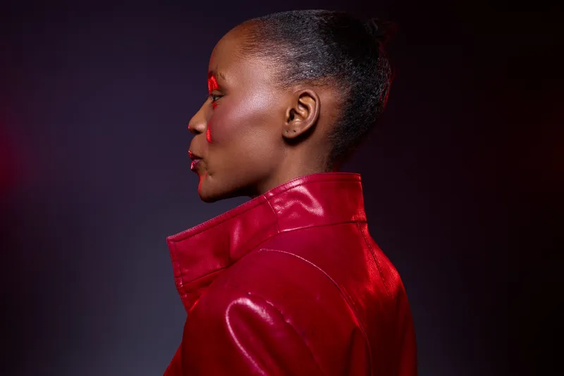 Profile of a serious African woman with dark skin wearing a shiny red leather jacket. Studio lighting highlights her smooth skin and elegant hairstyle on a dark background. Fashion concept.