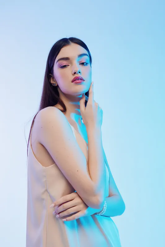 Young woman with long dark hair poses in a sleeveless dress under blue lighting. Her hand gently touches her neck, showcasing beauty and fashion concepts in a studio setting.