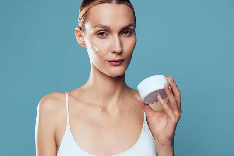 Young woman with short blonde hair applies face cream on her cheek while holding a white cosmetic jar against a blue background with a calm expression.