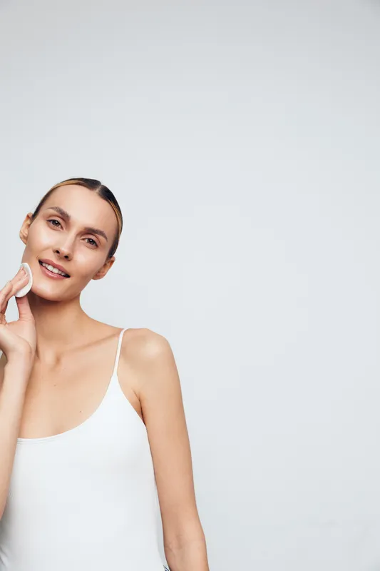 Young woman with clear skin gently cleansing her face with cotton pad, natural beauty routine, minimal makeup, white tank top, neutral background, skincare concept