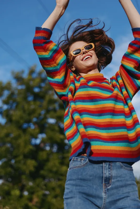 Woman in colorful striped sweater wearing sunglasses smile as she is jumping outdoors under blue sky. Casual jeans, joyful expression, motion and sunny lifestyle portrait.
