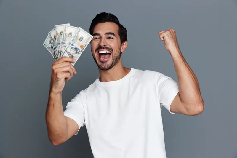 Happy cheerful man celebrating success while holding cash in hands, exuding joy and excitement, captured against a plain gray background. People lifestyle concept.