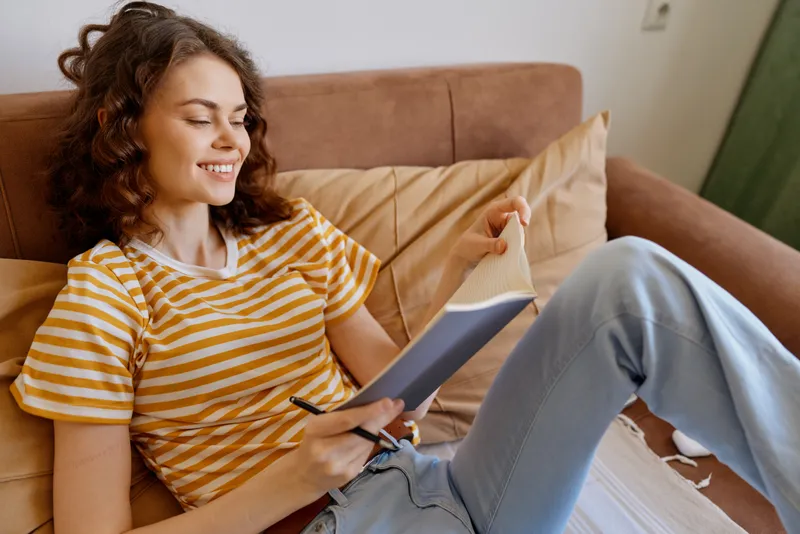 Calm woman relaxing on a sofa at home, writing in a notebook, wearing a striped shirt and jeans, warm daylight, casual lifestyle portrait in a cozy living room. Soft cushions around her create a