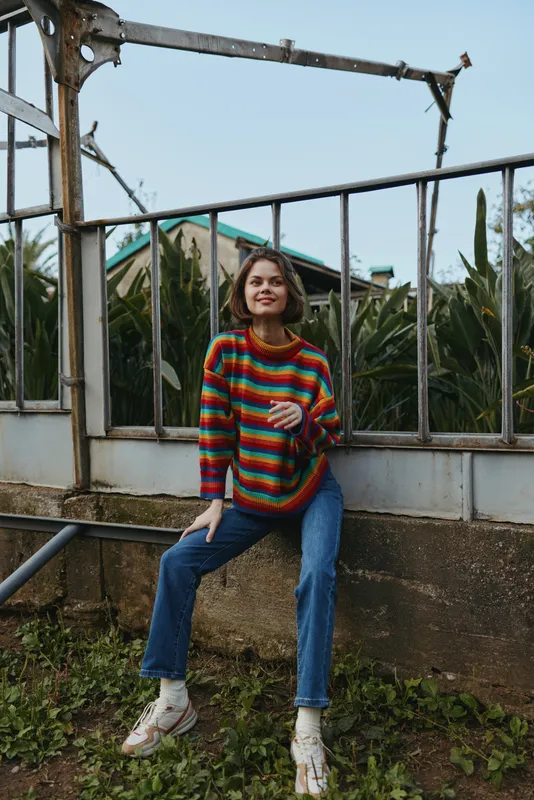 Young woman in a colorful striped sweater and casual outfit, blue jeans and white sneakers sitting on a concrete ledge by a greenhouse with tropical plants, smiling.