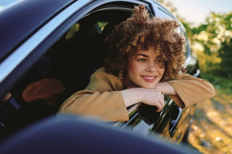 Smiling woman with curly hair leaning out of a car window, outdoor scene, casual jacket, relaxed pose. Retro film color and lifestyle vibe, fresh air, carefree moment.