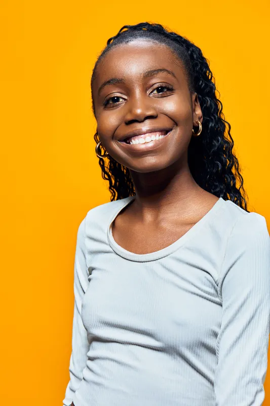 A cheerful young African girl with curly hair smiles brightly against a vibrant yellow background, showcasing her joyful expression and casual style