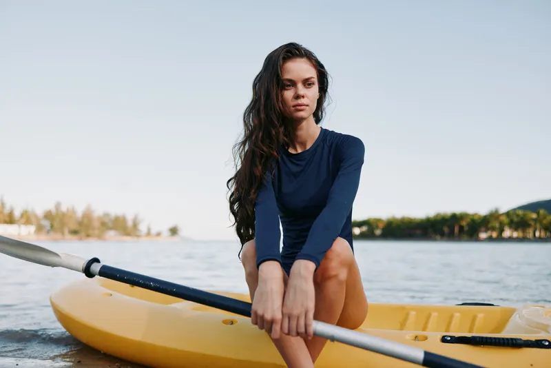 A young woman sits on a yellow kayak with a paddle across calm water, wearing a navy long sleeve top, gazing ahead with a thoughtful, athletic pose.