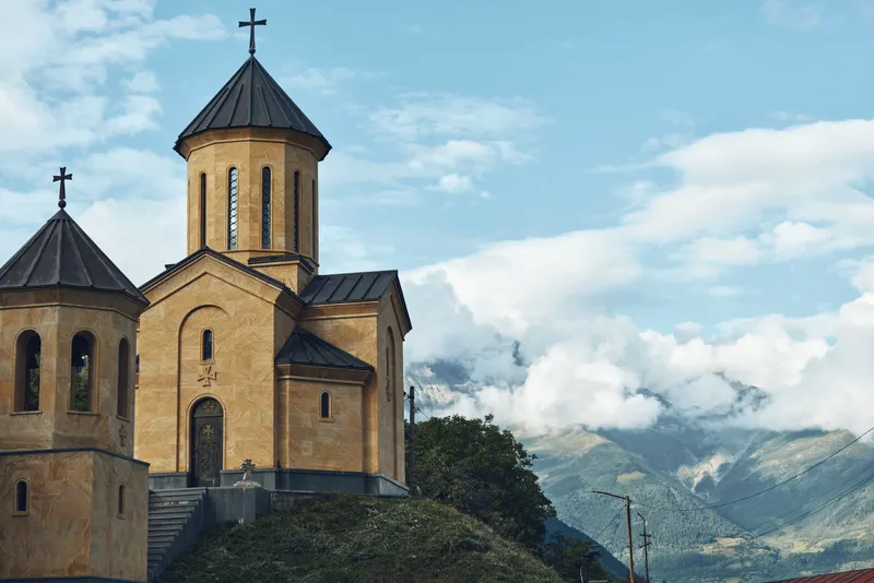 Church perched on a rugged hillside amid a dramatic mountain landscape, featuring domed towers and crosses, conveying serene religious architecture under a blue sky.