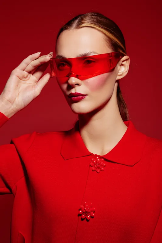 Stylish woman in red sweater and red translucent glasses poses against red background. Fashion portrait with modern accessories and bold color contrast for trendy editorial concept.