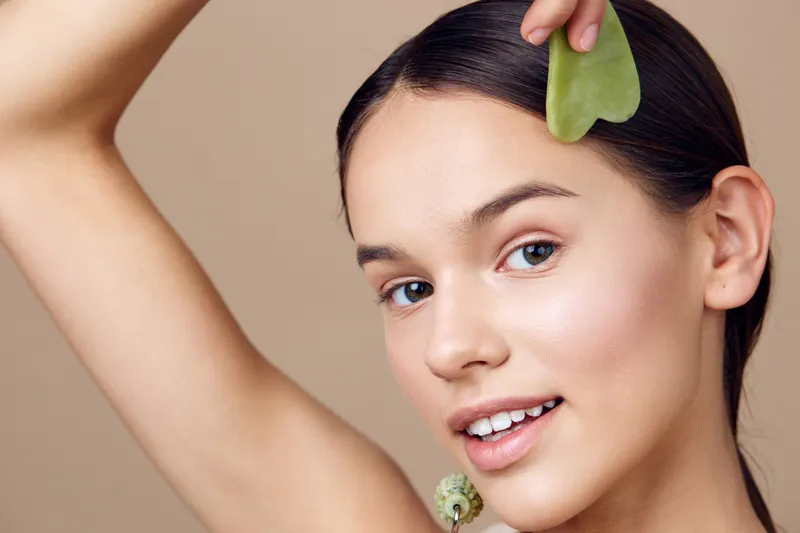 Young woman applying jade roller on her face, promoting healthy skin and beauty routine, with a serene expression against a neutral background