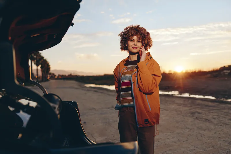 Woman lifestyle outdoors at sunset on road beside car door, casual jacket and jeans, smiling and standing, warm golden light, travel mood, street photography