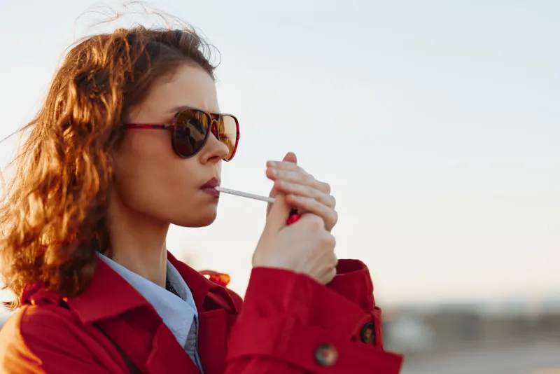 Young woman with curly hair wearing a red coat and sunglasses lights a cigarette outdoors during the day with a clear sky background.