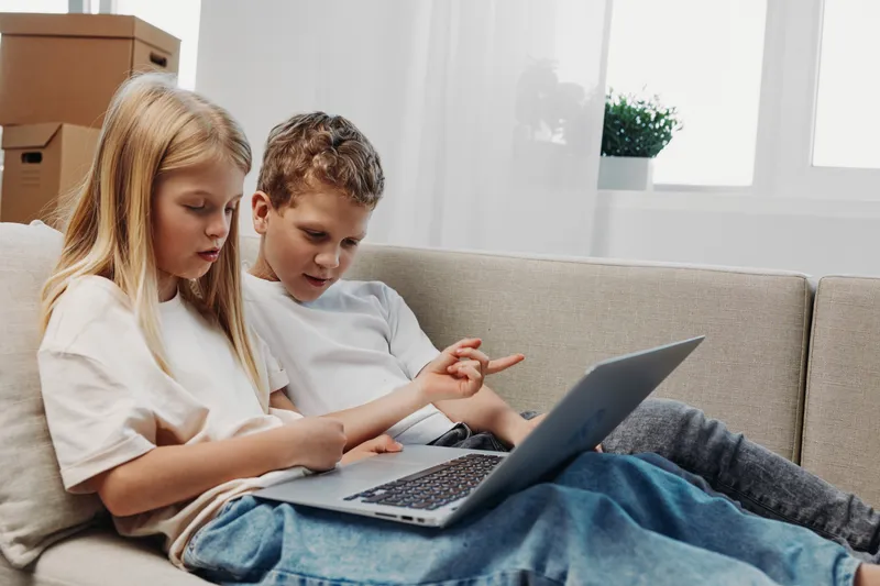 Two children sitting on a couch focused on a laptop screen, learning and exploring technology together in a bright living room environment.