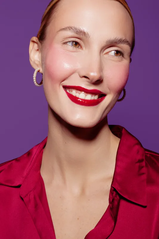 Smiling woman with red lipstick and hoop earrings wears a red blouse against purple background. Close-up portrait with natural skin and joyful expression. Beauty and fashion concept.