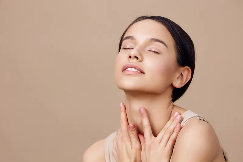 A serene young woman with a natural look gently caressing her neck, expressing tranquility against a soft beige background, highlighting beauty and wellness